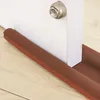 A close-up shot of a brown foam door draft stopper placed on a wooden floor against the bottom of a white door. Náhled obrázku produktu