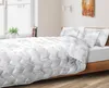 A bed with a white comforter and pillows with a floral pattern, set in a bedroom with a window and a plant. Náhled obrázku produktu