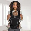 A woman is holding a baby in a black baby carrier. The background is a room with windows. Produktbild-Vorschau