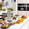 A kitchen counter with a breakfast spread including a juicer, fruits, pastries, and eggs. Produktbild-Vorschau