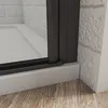 A close-up view of the bottom corner of a shower enclosure, showing the black frame, white base, and a brick-patterned wall. Produktbild-Vorschau