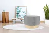 A modern living room setting with a grey and wood ottoman on a rug, next to a wooden stool and a framed artwork. Produktbild-Vorschau