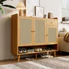 A wooden shoe cabinet with rattan doors and a drawer, filled with shoes, is placed in a room with a lamp and artwork on top. Voir les images du produit