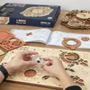 A person's hands are assembling a wooden 3D puzzle of a zodiac wall clock, with the box and instructions visible. Voir les images du produit