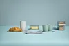A breakfast setting with a croissant, butter dish, two mugs, and a container of crackers on a light blue surface. Produktbild-Vorschau