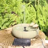 A solar-powered bird bath fountain with two decorative birds sits on a stone pedestal surrounded by lush green ferns. Voir les images du produit