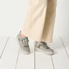 A person's legs wearing beige pants and grey sneakers, standing on a white wooden floor. Anteprima dell’immagine prodotto
