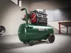 A green air compressor with a black and red motor unit is shown in a workshop or garage setting. Produktbild-Vorschau