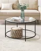 A round coffee table with a glass top and a woven basket underneath, placed on a rug in front of a sofa. Náhled obrázku produktu