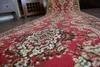 A close-up, low-angle view of a red carpet with a traditional pattern, unrolled on a wooden floor. Furniture is visible in the background. Produktbild-Vorschau