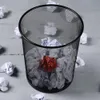 A black metal mesh trash can filled with crumpled paper, with one red crumpled paper ball prominently placed in the center. Several crumpled white paper balls are scattered around the trash can on a light gray wooden surface. Náhled obrázku produktu