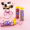 A collection of decorative sprinkles and marshmallow pops arranged on a pink surface. Produktbild-Vorschau