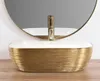 A close-up view of a gold and white washbasin with a gold faucet, placed on a wooden surface, with a round mirror in the background. Náhľad obrázku produktu