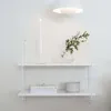 A white wall with a white shelf mounted on it. The shelf holds a candle, a vase with greenery, a stack of books, and a round container. Produktbild-Vorschau