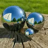 Three polished stainless steel garden spheres of different sizes are placed on a weathered wooden surface, with a green grassy background and a building reflected in the spheres. Náhled obrázku produktu