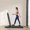 A woman is walking on a treadmill in a room with white paneled walls and a window with gray curtains. Náhled obrázku produktu