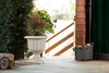 A cream-colored planter with plants sits on a wooden deck next to a wooden structure and a pile of logs. Voir les images du produit