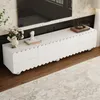 A white TV stand with drawers and doors is shown in a living room setting, with a TV mounted on the wall above it. Decorative items like a vase with flowers and a small sculpture are placed on top of the stand. A rug is on the floor in front of the stand. Produktbild-Vorschau
