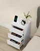A white nightstand with three drawers, next to a bed. The top of the nightstand has a phone charging station and an alarm clock. Náhled obrázku produktu