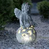 An angel statue with internal lighting sits on gravel in a garden setting. Produktbild-Vorschau