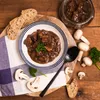 A bowl of pork goulash with mushrooms and bread slices on a wooden table. Produktbild-Vorschau