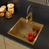 A modern kitchen sink with a gold finish, a black faucet, and three red tomatoes inside. Jars of nuts are visible on the counter. Produktbild-Vorschau