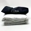 A folded white weighted blanket is stacked on top of another folded white weighted blanket. A black fabric bag with the white text "CURA OF SWEDEN" is placed on top of the blankets. Podgląd zdjęcia produktu
