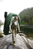 A man wearing fishing gear and carrying an inflatable boat on his back stands on rocks by a lake, with a forest-covered mountain in the background. Podgląd zdjęcia produktu