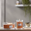 Two lit candles and a decorative bowl on a wooden surface, with a blurred background of a shelf and hanging plant. Produktbild-Vorschau