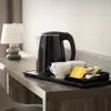 A black electric kettle, two white cups, and tea packets are arranged on a wooden desk next to a small lamp. Náhled obrázku produktu