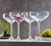 Four clear champagne glasses are arranged on a white surface against a gray textured background. One glass is filled with a pink beverage, and a bunch of green grapes is visible to the right. Produktbild-Vorschau