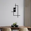 A modern black metal wall clock with an abstract geometric design is mounted on a light gray wall above a wooden dining table with decorative plants and vases. Anteprima dell’immagine prodotto
