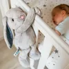 A plush bunny toy hangs on the side of a crib next to a sleeping baby. Produktbild-Vorschau