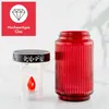 A red glass jar and a clear LED candle lantern are displayed on a white surface with a light gray marble pattern background. A red graphic overlay with a diamond icon and German text is in the top left corner. Produktbild-Vorschau