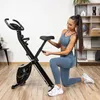 A woman is adjusting the seat of a black foldable exercise bike in a home gym setting. Náhľad obrázku produktu