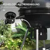 A close-up marketing image of a black charcoal grill with a small inset showing an ash catcher. The background features lush green plants and foliage. Produktbild-Vorschau