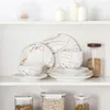 A set of white and gold marble-patterned porcelain dishes is neatly arranged on a white shelf. Produktbild-Vorschau