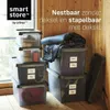Several storage boxes are stacked and arranged in a room with wooden walls and flooring. One box is filled with blankets and clothes, while others contain various items. Produktbild-Vorschau