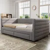 A grey upholstered daybed with two drawers is shown in a well-lit room with large windows. Produktbild-Vorschau