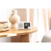 A white Blaupunkt portable radio sits on a wooden table next to a decorative vase and a candle. The scene is lit by natural light coming from a window with sheer curtains. Náhled obrázku produktu