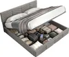 A bed with its storage compartment open, showing various items stored inside. The bed has a gray upholstered headboard and frame, and is made up with white bedding and a gray throw blanket. Voir les images du produit