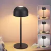 A black, modern LED table lamp with a dome-shaped shade is displayed on a wooden surface, possibly a nightstand. The lamp is lit, casting a warm glow. The image also includes smaller inset images showing the lamp in different lighting conditions. Náhľad obrázku produktu