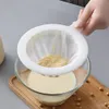 A hand is holding a white mesh strainer over a glass bowl filled with a light-colored liquid. The strainer contains a yellow, pulpy substance. Soybeans are scattered on a wooden board beneath the bowl. Produktbild-Vorschau