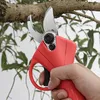 A hand holding a red electric pruning shear to cut a tree branch. Náhled obrázku produktu