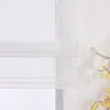A close-up view of a white sheer Roman blind with horizontal bands and loops, partially covering a window. A decorative branch with light-colored leaves is visible to the right. Produktbild-Vorschau