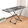 A black metal drying rack is set up on a wooden floor in front of a wall with floral wallpaper. A small white planter with green plants is to the left of the drying rack. Produktbild-Vorschau