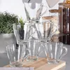 A stack of clear glass mugs on a wooden surface, with a plant and jars in the background. Anteprima dell’immagine prodotto