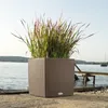 A brown woven planter with tall green and red ornamental grass is placed on a concrete surface next to a body of water with a wooden pier and a sailboat in the distance. Produktbild-Vorschau