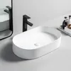 A white oval countertop sink with a black faucet is shown on a light gray surface, with a mirror in the background and some toiletries on a tray to the right. Náhľad obrázku produktu