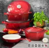 A collection of red enameled cast iron cookware, including pots and a frying pan, arranged on a countertop with some fresh vegetables and herbs. Voir les images du produit